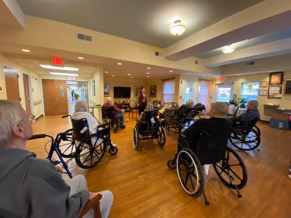 A group of elderly residents in wheelchairs gathered in a communal living room while a staff member stands near the front.