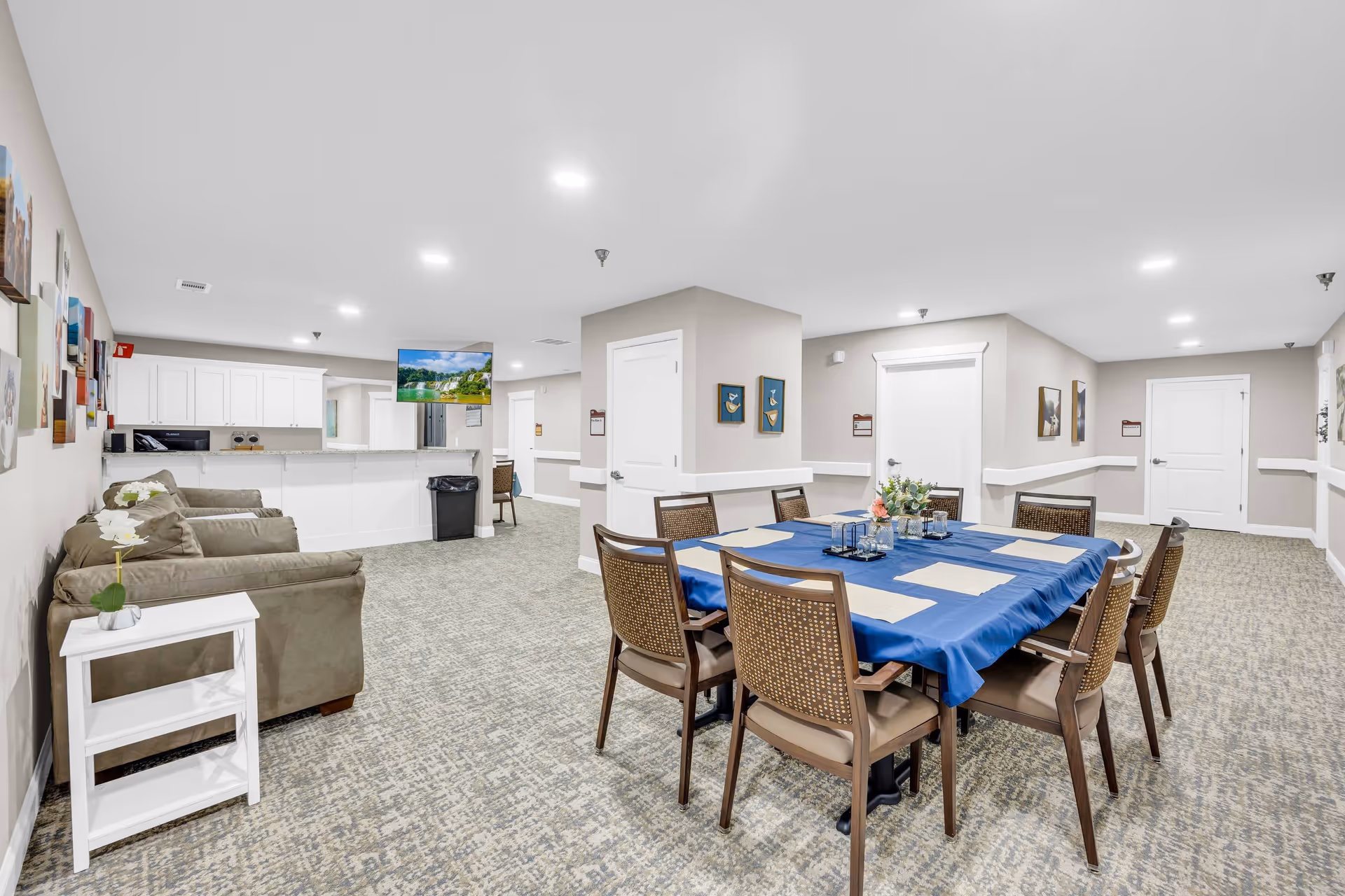 A bright and spacious common area in a senior living facility featuring a dining table with eight chairs covered with a blue tablecloth and placemats. To the left, there are two cushioned armchairs next to a small white side table with a potted orchid. The background shows a kitchen area with white cabinets and a granite countertop, and a wall-mounted TV displaying a nature scene. The walls are decorated with framed pictures and the floor is carpeted with a patterned design.