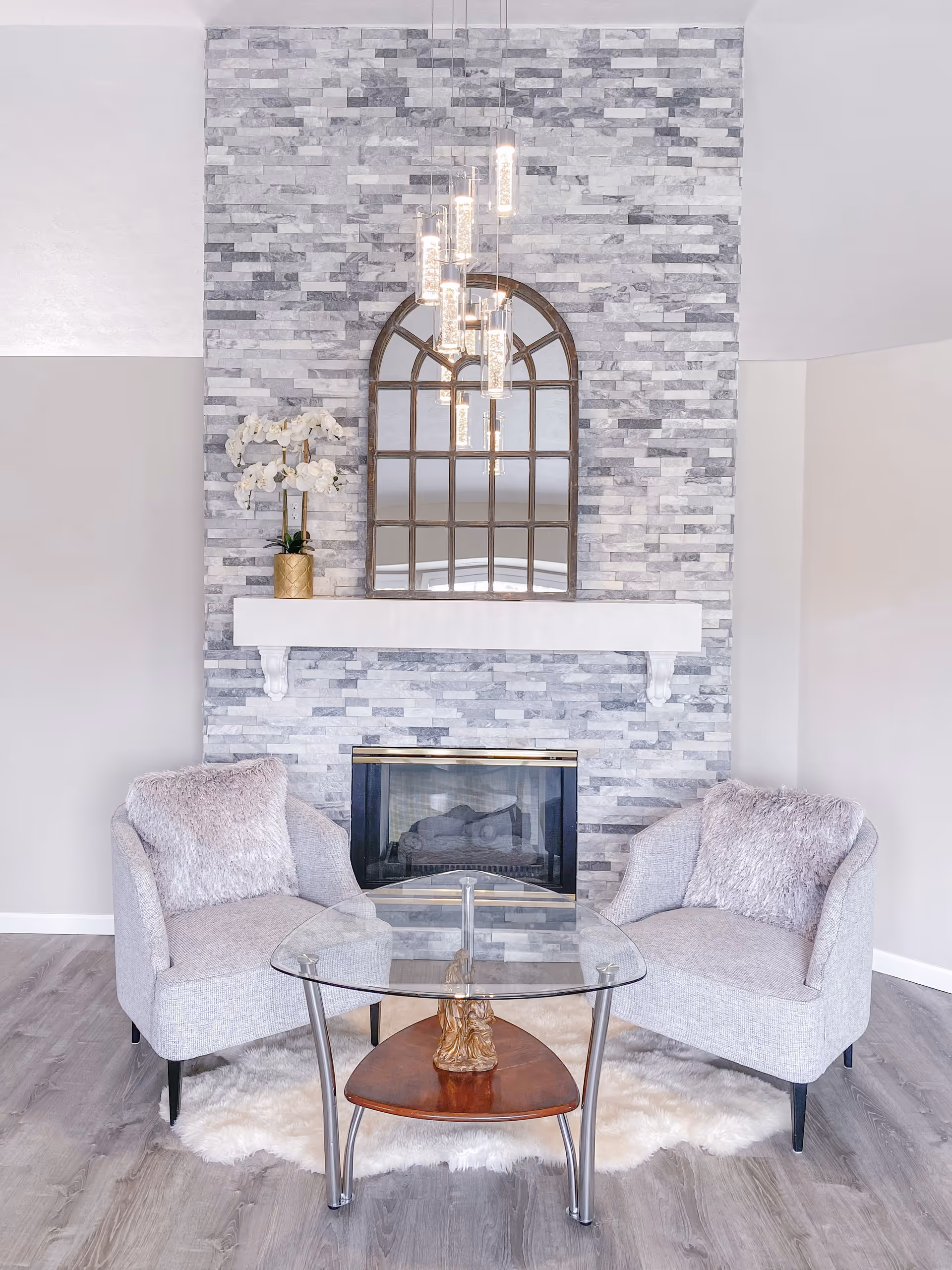 A cozy living room area featuring two light gray armchairs with fluffy cushions placed on a white fur rug. Between the chairs is a glass coffee table with a wooden lower shelf. Behind the chairs is a modern fireplace with a gray stone brick wall extending to the ceiling. Above the fireplace is a white mantel holding a gold vase with white orchids and a large arched window-style mirror. A contemporary chandelier with multiple hanging lights is reflected in the mirror.