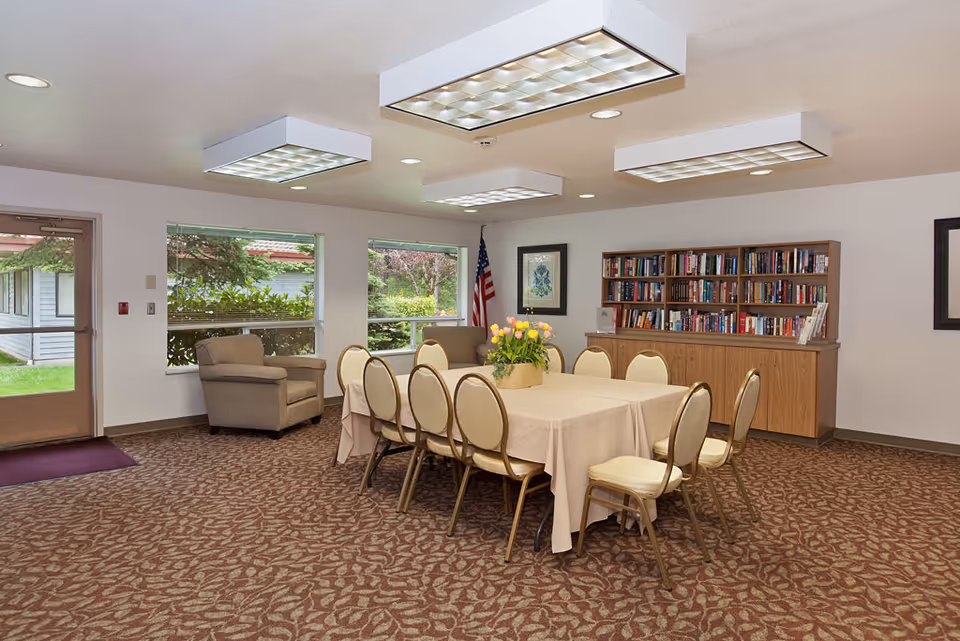 Well-lit communal dining/activity room with a table and chairs, bookshelves, armchairs, and windows looking out to greenery.