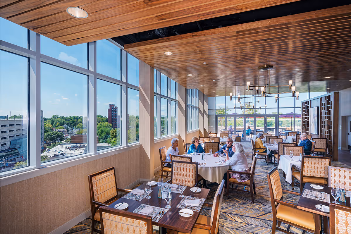 Bright, upscale dining room with large windows overlooking the city and seniors seated at tables.