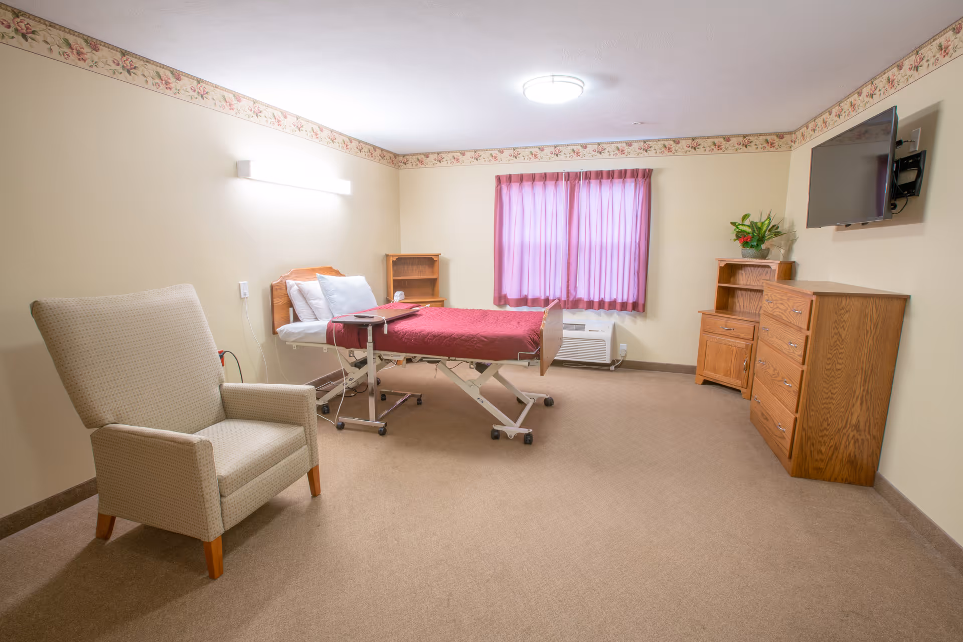 A nursing facility room with a hospital bed covered in a red blanket, a beige armchair, wooden furniture including a dresser and a small bookshelf, a window with pink curtains, and a wall-mounted TV.