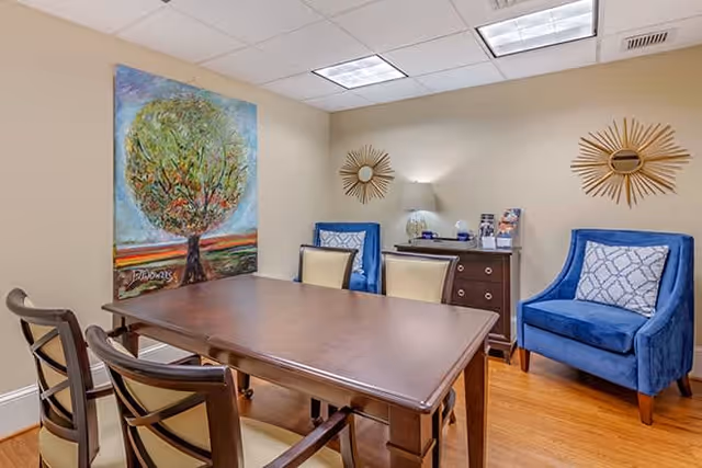 A small meeting or sitting room with a wooden table surrounded by four chairs, two with beige cushions and two blue upholstered armchairs with patterned pillows. The room has beige walls, a large colorful painting of a tree on one wall, two sunburst mirrors, a small wooden cabinet with a lamp and decorative items, and a hardwood floor.