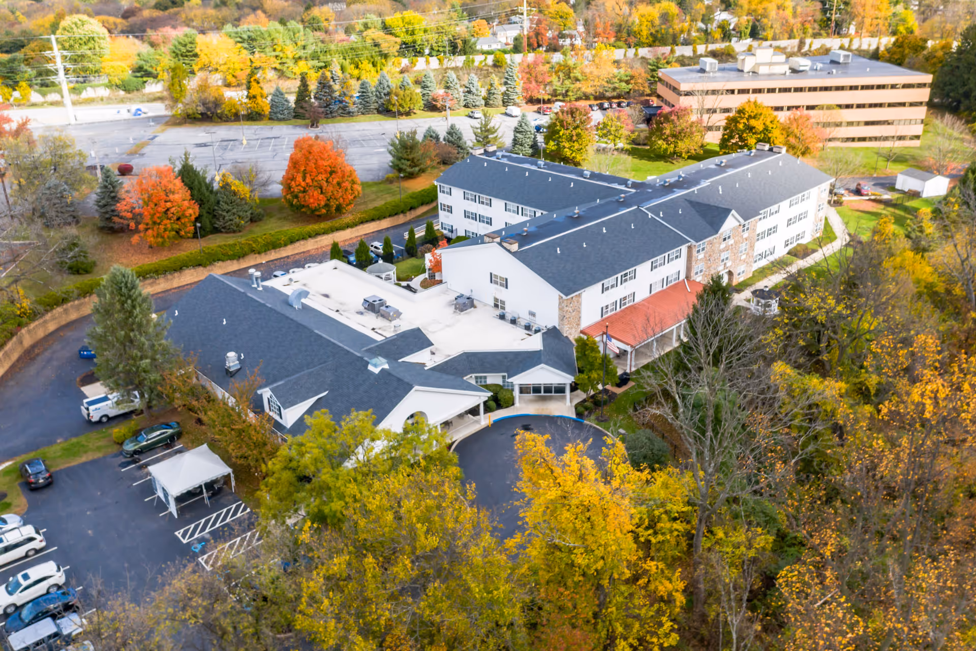 Aerial view of Rittenhouse Village At Lehigh Valley, showing a large senior living facility building surrounded by trees with autumn foliage, parking lots, and a nearby office building.