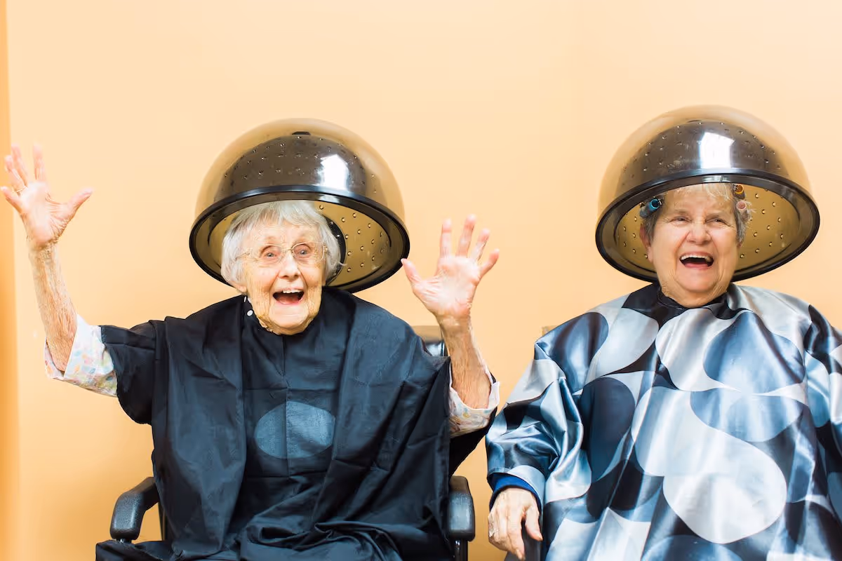 Two elderly women sitting side by side under hair drying machines in a salon setting, both smiling and appearing joyful. One woman has her hands raised excitedly, and both are wearing salon capes.