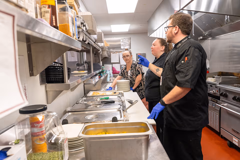 Three kitchen staff members wearing gloves and uniforms are working in a commercial kitchen. They are standing near a counter with food trays and plates, with stainless steel appliances and shelves in the background.