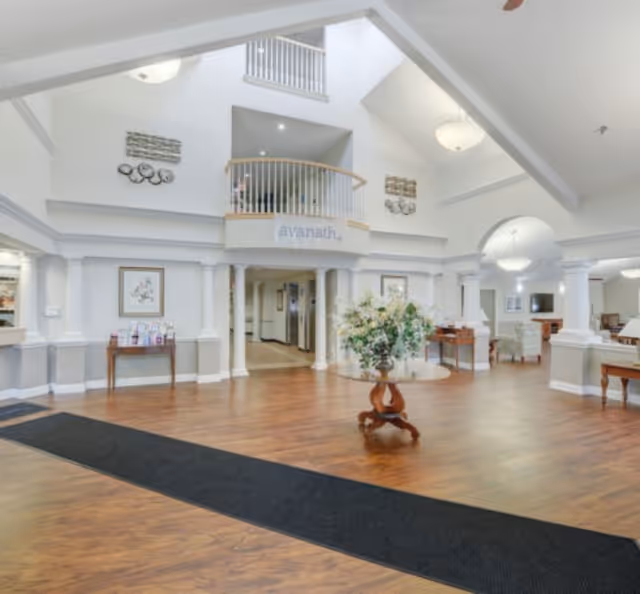 Spacious senior living lobby with a vaulted ceiling, central floral table, wood floors, and an upper balcony overlooking the entrance.