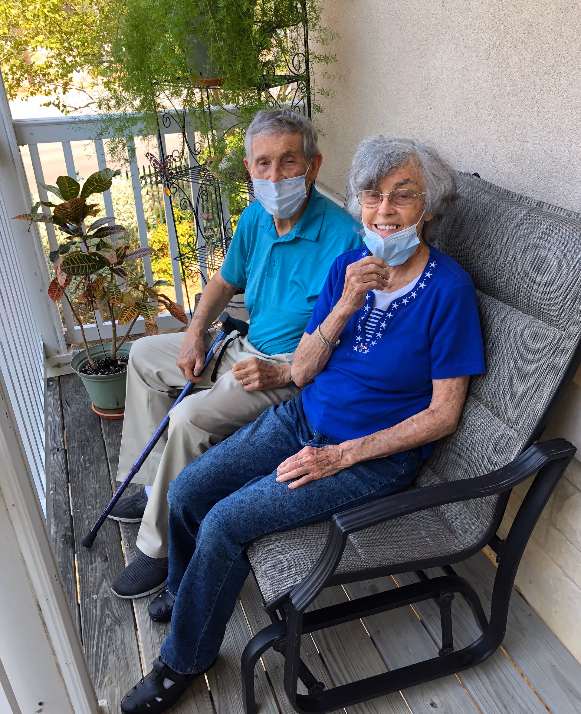 An elderly man and woman wearing face masks sit on a cushioned outdoor bench on a wooden porch. The man is holding a cane and wearing a turquoise polo shirt and beige pants, while the woman is dressed in a blue shirt and jeans. There are potted plants and a decorative metal plant stand behind them.
