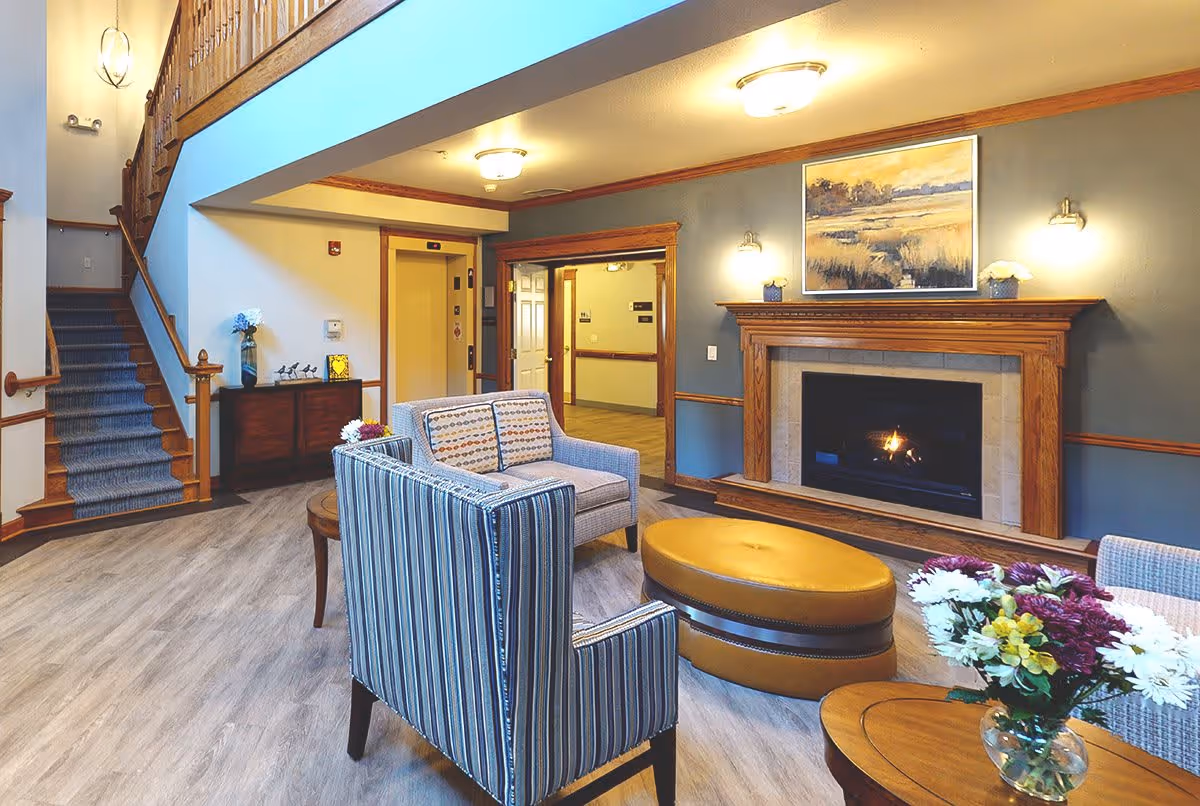 A cozy living room area in a senior living facility with a fireplace, two striped armchairs, a loveseat, a round yellow ottoman, and a wooden coffee table with a vase of flowers. The room features wood trim, a staircase with blue carpeting, an elevator, and a painting above the fireplace.
