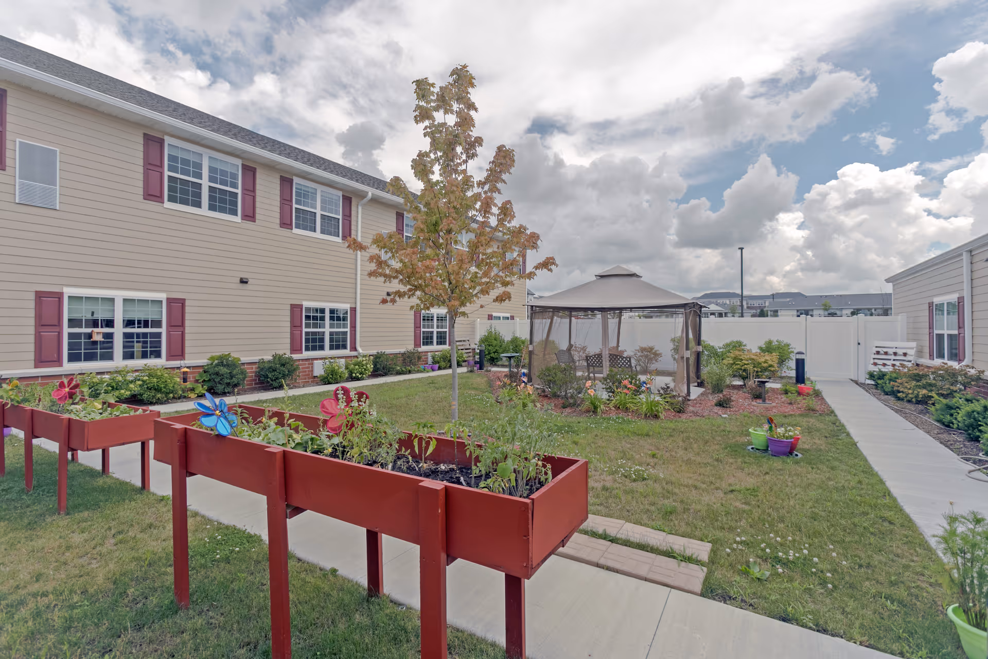 Outdoor garden area at a senior living facility with raised red planter boxes containing plants and decorative butterflies, a small tree in the center, a gazebo with seating, and beige buildings with maroon shutters surrounding the garden under a partly cloudy sky.