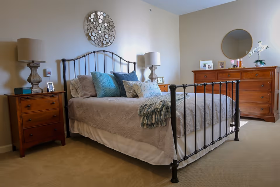 A cozy bedroom with a metal frame bed adorned with multiple pillows and a throw blanket. On either side of the bed are wooden nightstands with matching lamps. A wooden dresser with a round mirror and decorative items is against the wall. The room has beige walls and carpeted flooring.