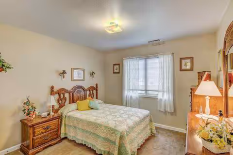 Well-lit traditional furnished bedroom with a double bed, wooden headboard, matching nightstands and a dresser by a window with sheer curtains.