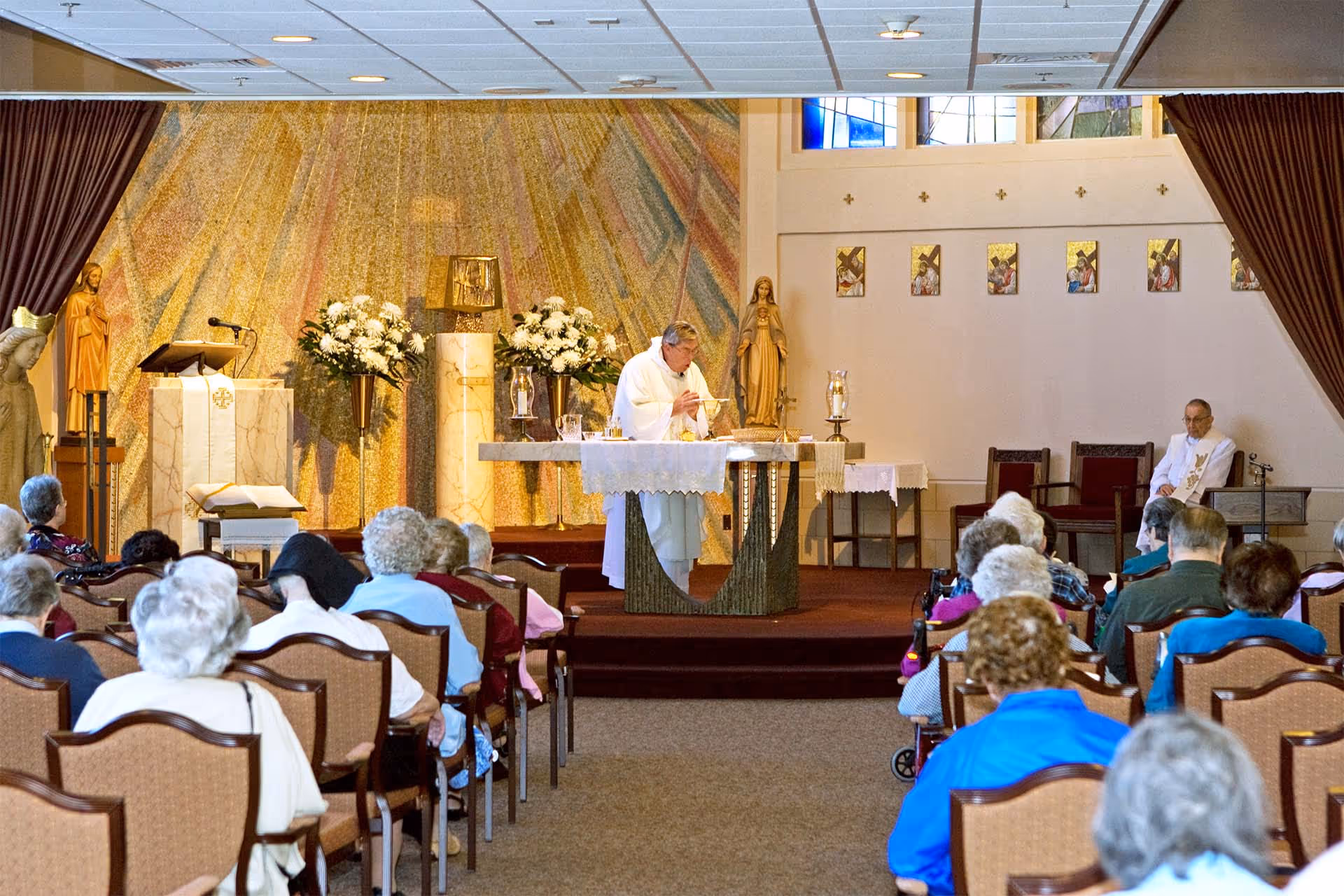 Residents seated in chairs attend a religious service in a chapel while a priest stands at the altar.