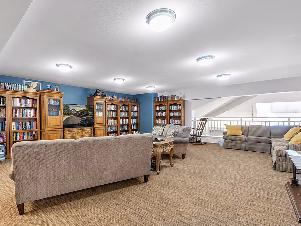 A spacious living room area with multiple sofas and armchairs arranged around a wooden coffee table. The back wall features several wooden bookshelves filled with books and a flat-screen TV in the center. The room has beige carpeting, white ceiling with multiple ceiling lights, and a blue accent wall behind the bookshelves. There is also a wooden rocking chair near a white railing overlooking a lower level.