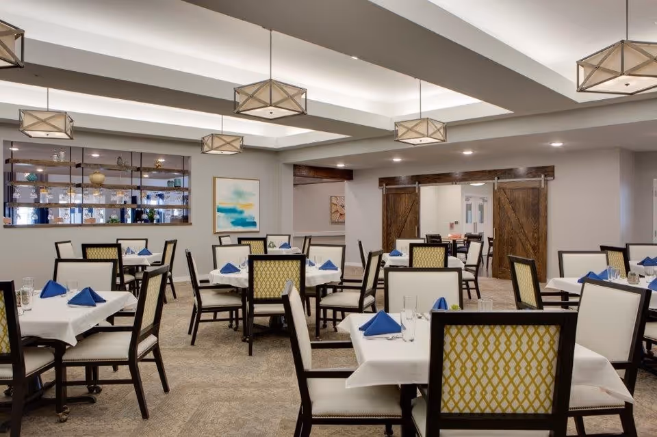 A dining room with multiple tables covered in white tablecloths, each set with blue folded napkins and glassware. The chairs have white cushions with a yellow patterned backrest. The room features modern pendant lights hanging from the ceiling, a large window with shelves displaying decorative items, and wooden sliding barn doors in the background.