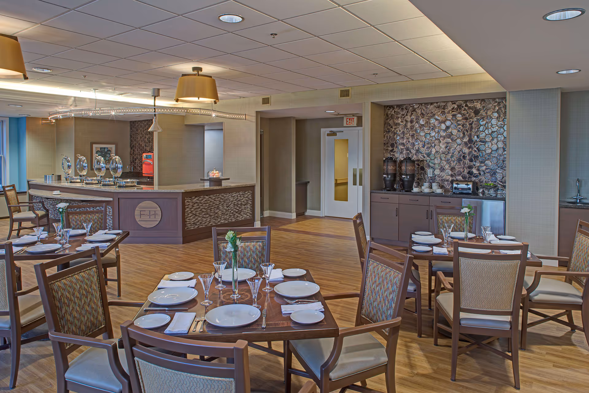 A dining room in a senior living facility with several wooden tables set with white plates, glasses, and silverware. The room features wooden flooring, cushioned chairs, a buffet counter with decorative elements, and a beverage station with coffee urns and cups against a textured wall. The ceiling has recessed lighting and hanging lamps.