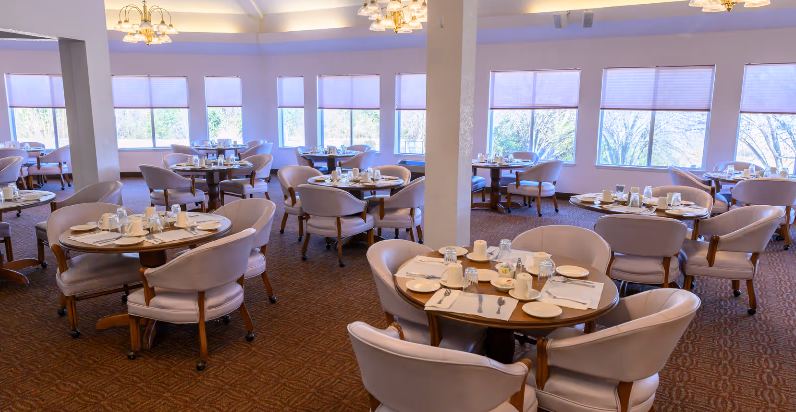 A spacious dining room with multiple round tables set for meals, each table surrounded by cushioned chairs. Large windows with blinds allow natural light to fill the room, and elegant chandeliers hang from the ceiling.