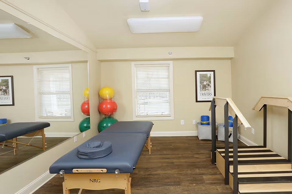 A therapy or exercise room with a padded treatment table, a large wall mirror, colorful exercise balls stacked in a corner, and a wooden parallel bar setup for walking or balance exercises. The room has light-colored walls, a window with blinds, and framed artwork on the wall.