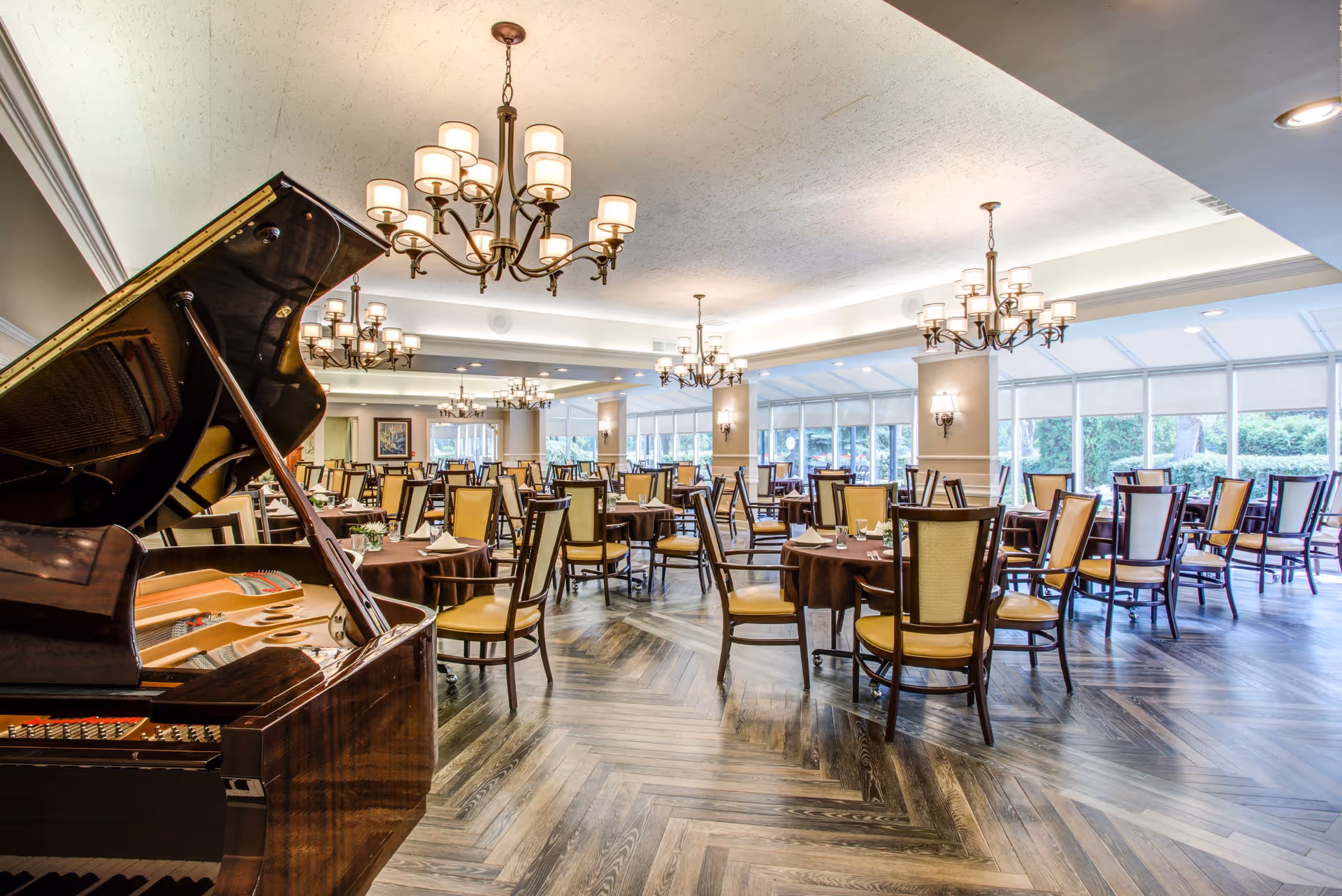 A spacious dining room with multiple round tables covered with brown tablecloths and surrounded by wooden chairs with yellow cushions. A grand piano is positioned on the left side of the room. The room is well-lit with several elegant chandeliers hanging from the ceiling and large windows along the right wall allowing natural light to enter. The floor has a herringbone wood pattern.