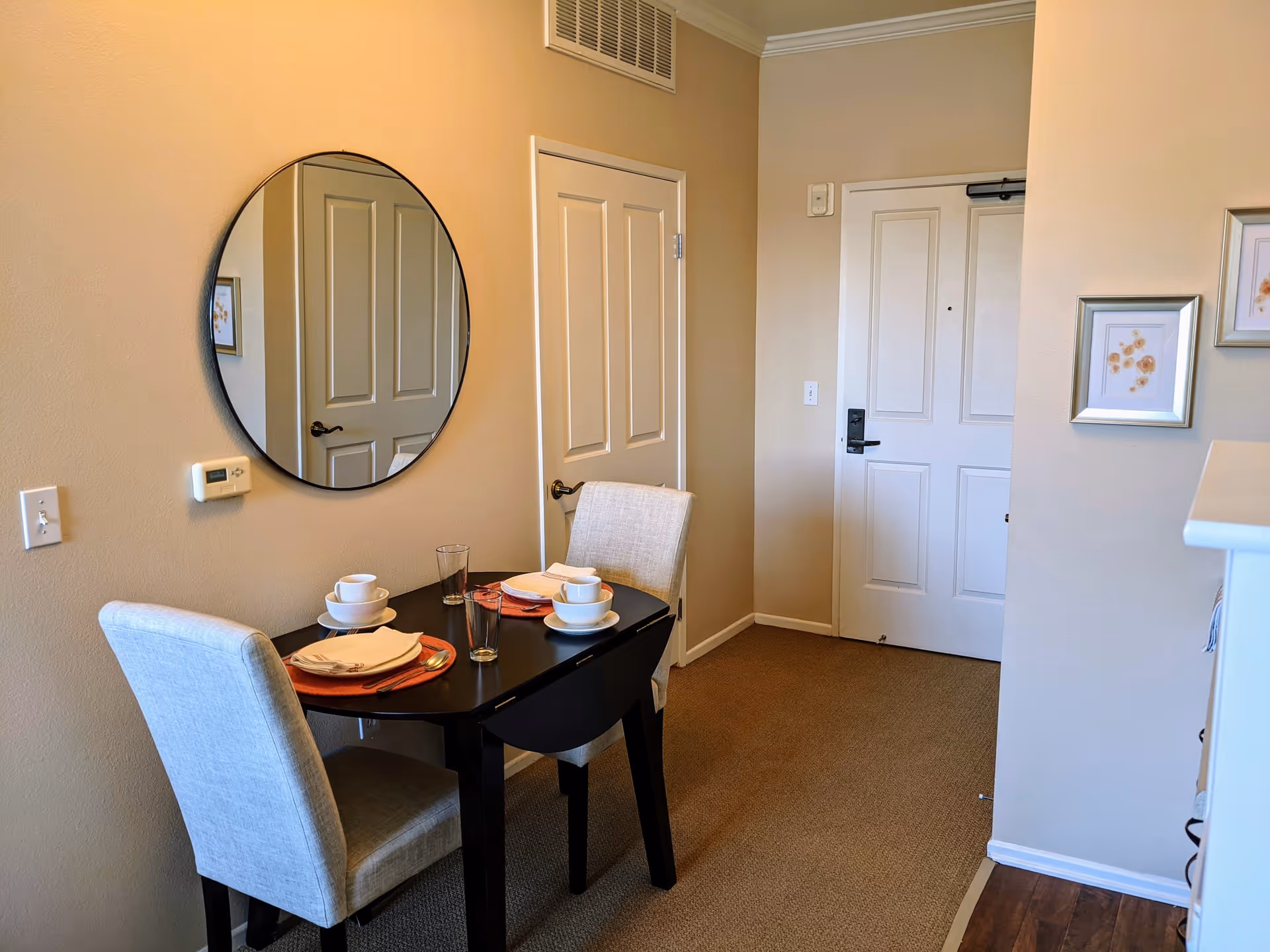 Small dining area with a black table set for two with white cups, plates, and glasses. Two upholstered chairs are positioned at the table. A round mirror hangs on the beige wall above the table. There are two closed white doors and a white entrance door in the background. The floor is carpeted except for a small section of wood flooring on the right.