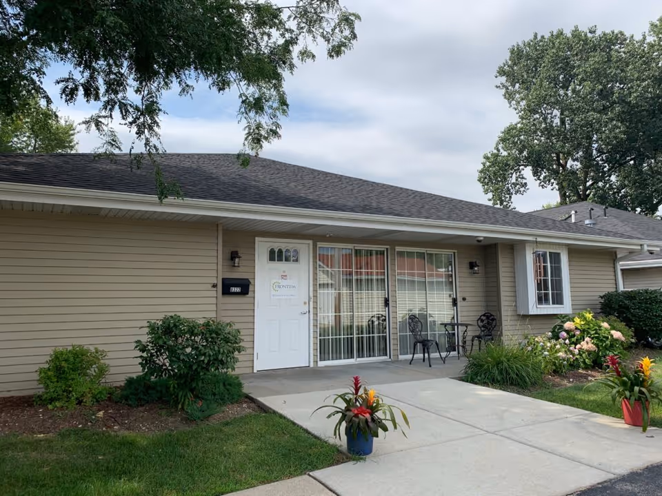 Single-story beige assisted living building front showing a white entry door, sliding glass doors with a small patio set, and potted flowers on the walkway.