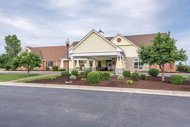 Exterior view of a single-story senior living facility building with a covered entrance, surrounded by neatly trimmed bushes, small trees, and a paved driveway under a cloudy sky.