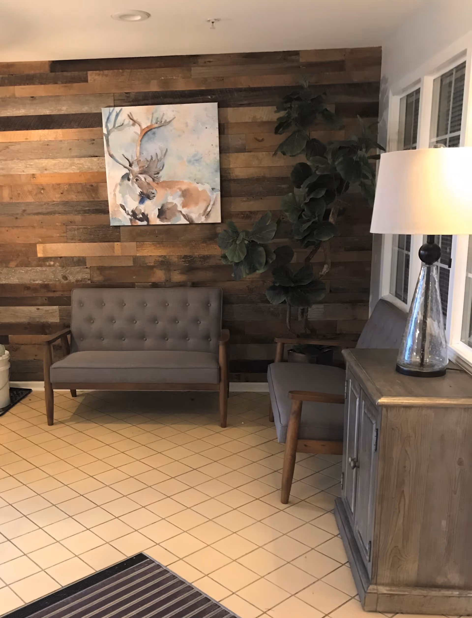A cozy seating area with two gray cushioned wooden armchairs and a wooden cabinet with a glass lamp on top. The background features a rustic wooden plank wall with a painting of a deer and a large green potted plant next to the chairs. The floor is tiled with light beige square tiles.
