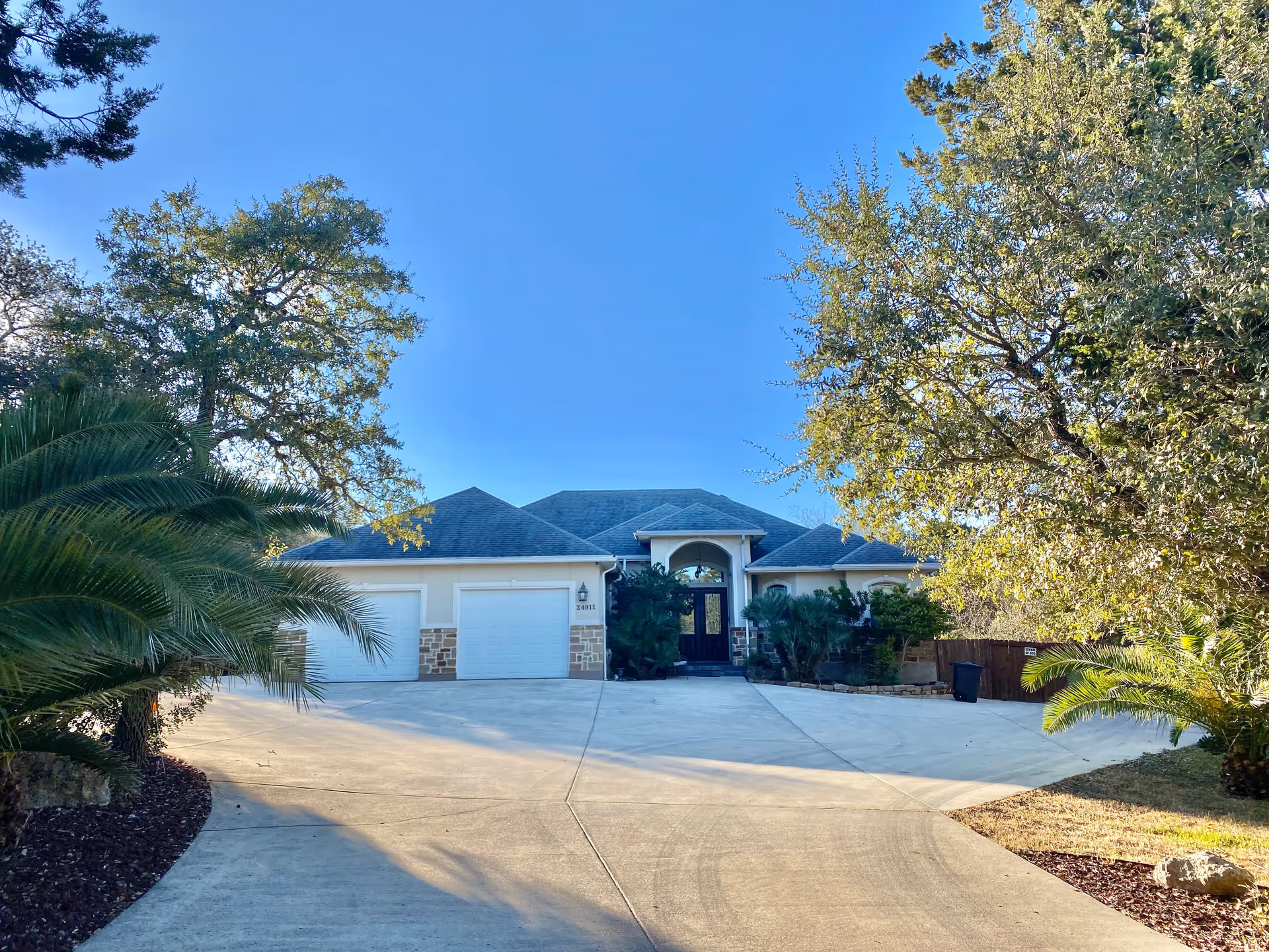 Front exterior view of a single-story residential building with a three-car garage, surrounded by trees and landscaping under a clear blue sky.
