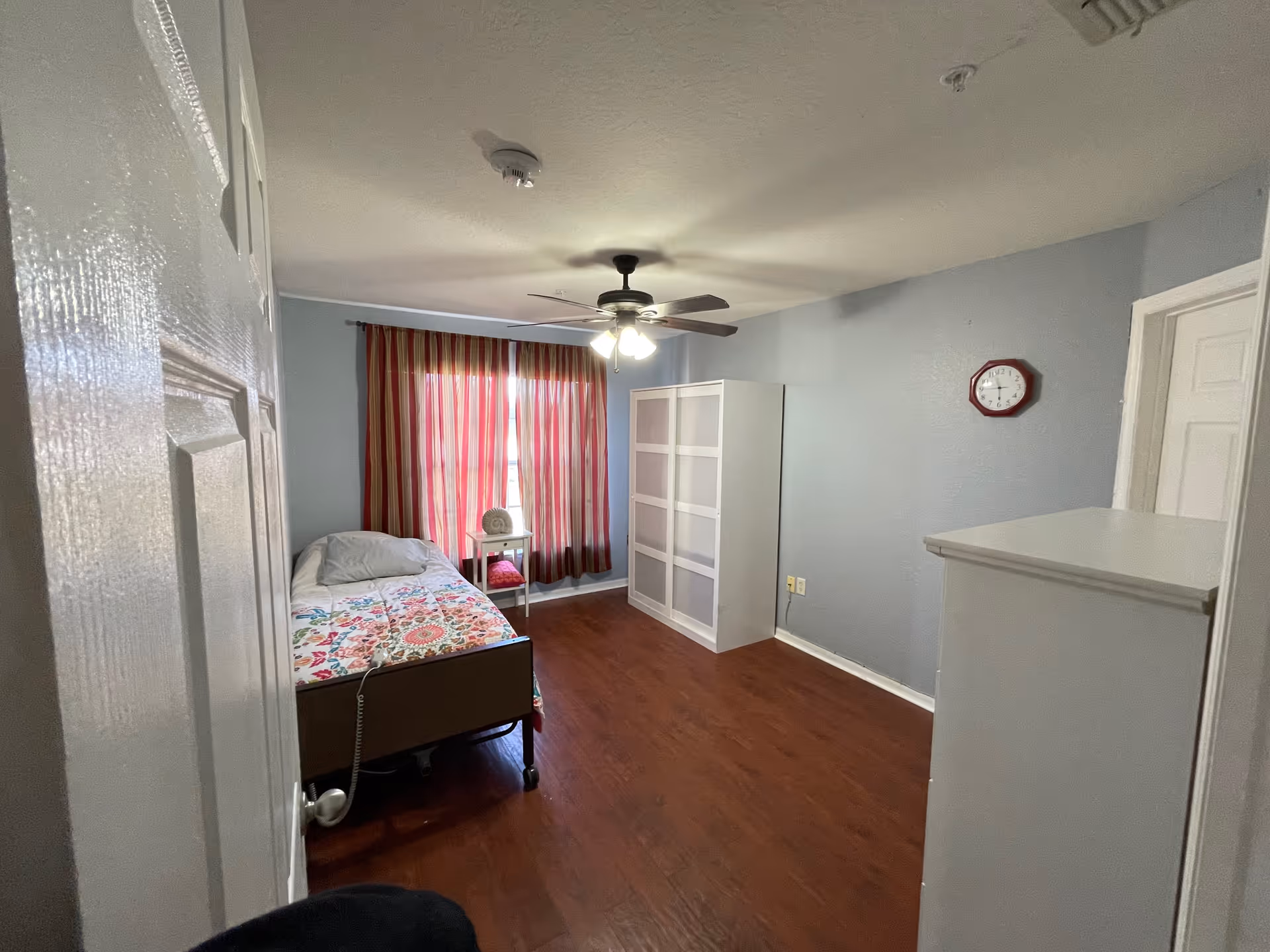 A small bedroom in an assisted living facility with a single bed covered with a colorful floral blanket, a white pillow, and a white side table next to the window with red and beige striped curtains. The room has light blue walls, a ceiling fan with lights, a white wardrobe, a white dresser, and a red wall clock. The floor is wooden.