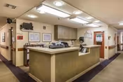 Interior view of a reception or nurse station area in a senior living facility with a large counter, overhead fluorescent lighting, wall-mounted TV, framed pictures on the walls, and a hallway leading to other rooms.
