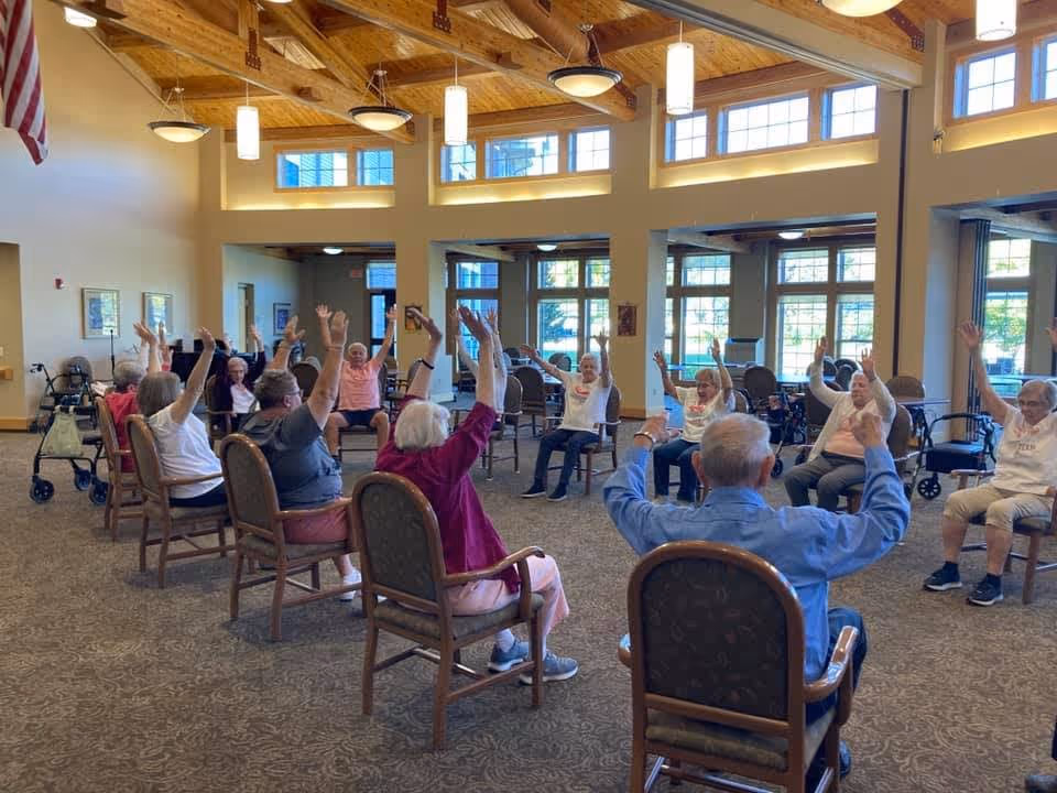A group of elderly individuals seated in a spacious, well-lit room with high ceilings and large windows, participating in a seated exercise or stretching activity with their arms raised. The room has wooden beams on the ceiling and comfortable chairs arranged in a circle.