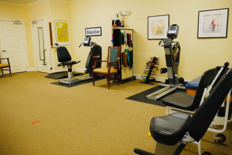 A senior living facility exercise room with two stationary exercise bikes, chairs, a small rack with exercise bands, and framed artwork on the walls. The room has beige walls and carpeted floor.