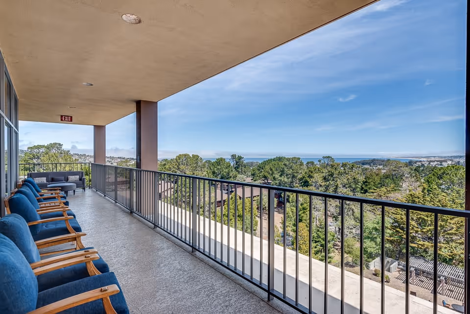 A spacious outdoor balcony at Lake Park Senior Living with several blue cushioned wooden chairs lined up along the left side and a small seating area with a sofa and table at the far end. The balcony overlooks a scenic view of trees, rooftops, and a distant body of water under a clear blue sky.