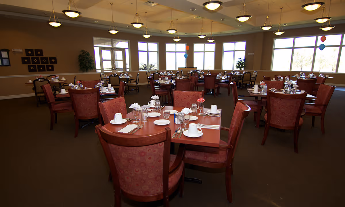 A spacious dining room in a senior living facility with multiple tables set for meals. Each table has red cushioned chairs, white plates, cups, glasses, and silverware neatly arranged. The room is well-lit with large windows allowing natural light to enter and ceiling lights providing additional illumination. There are some decorative plants and wall art visible in the background.