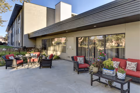 Outdoor patio area at Winslow Court Assisted & Senior Living with wicker chairs and sofas featuring red cushions and patterned pillows, a small round table with plants, and a rectangular coffee table with various potted plants. The patio is adjacent to a beige building with large windows and sliding glass doors.