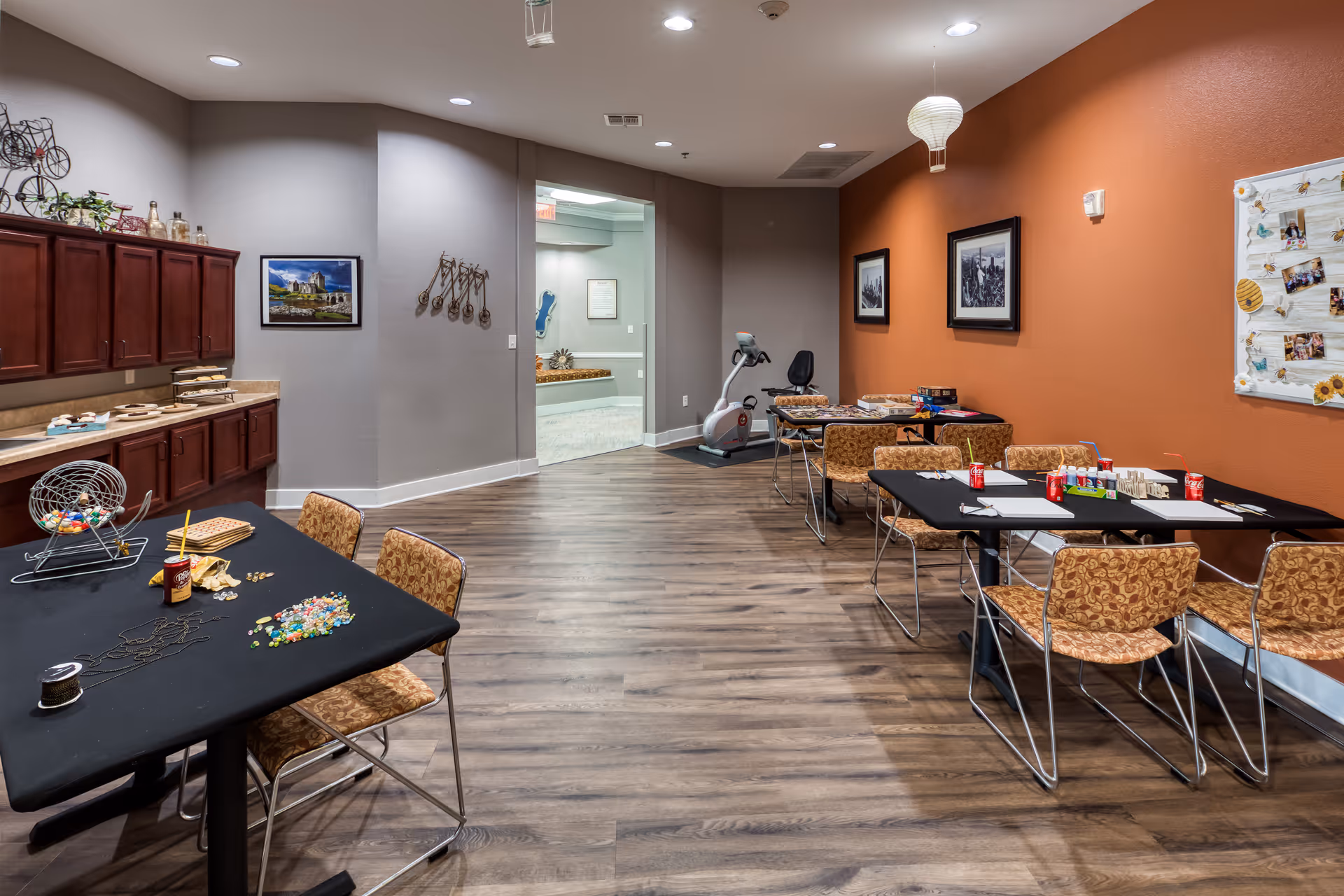A spacious interior activity room with tables and chairs, craft supplies on the tables, cabinets along the left wall, and an exercise bike toward the back.