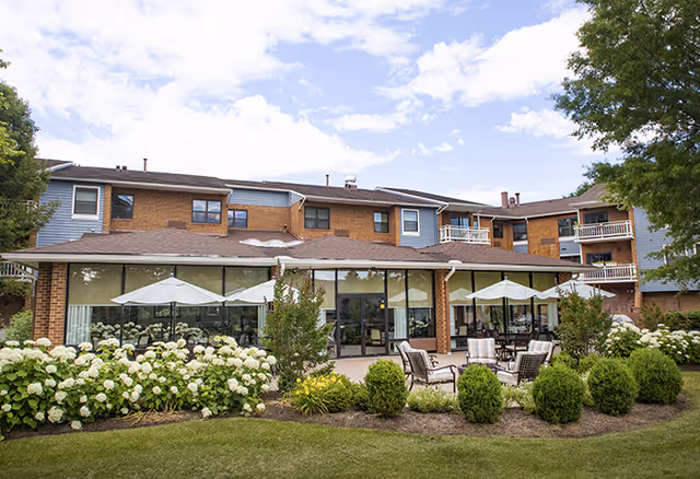 Exterior view of AVIVA Baldwin Park senior living facility showing a two-story building with large windows, outdoor seating area with chairs and tables under umbrellas, surrounded by well-maintained landscaping including bushes, flowers, and trees under a partly cloudy sky.