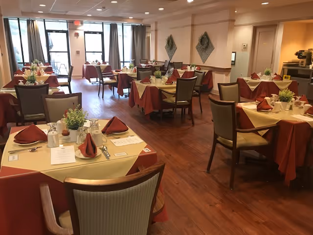 A dining room in Regency Park Senior Living Community with multiple tables set for meals. Each table has a yellow tablecloth with a red overlay, neatly folded red napkins, menus, and small green plants as centerpieces. The room has wooden flooring, chairs with cushioned seats, and large windows with curtains allowing natural light to enter.