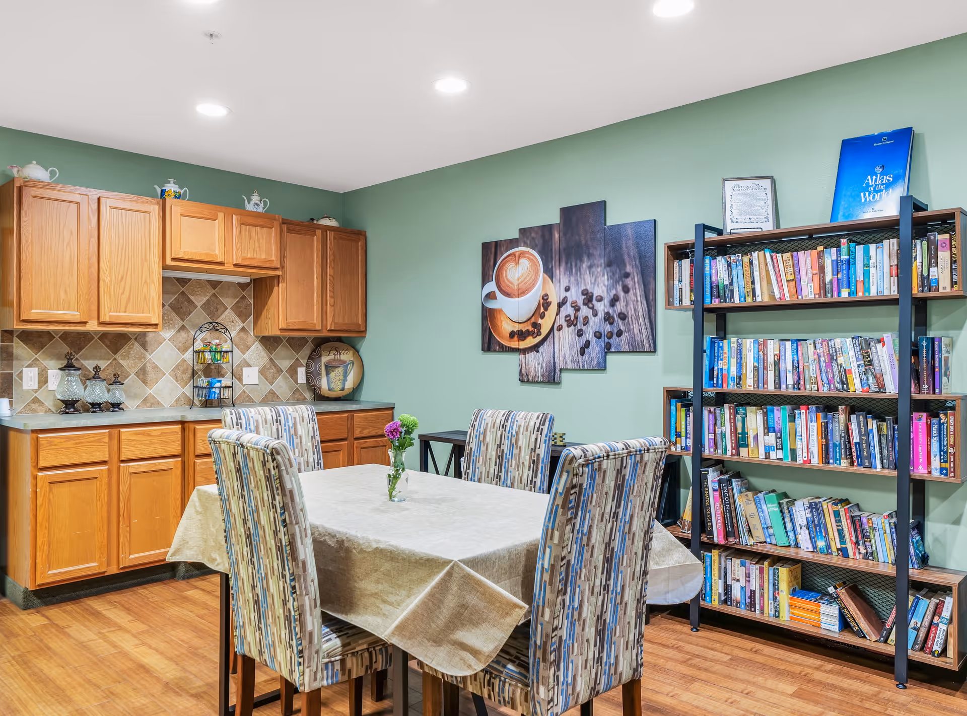 A cozy dining area in an assisted living facility featuring a table with a beige tablecloth and four patterned chairs. Behind the table is a wooden cabinet with a tiled backsplash and decorative items on the countertop and above the cabinets. On the right side, there is a tall bookshelf filled with books, and a wall art piece depicting a cup of coffee with coffee beans on a wooden surface hangs above a small side table.