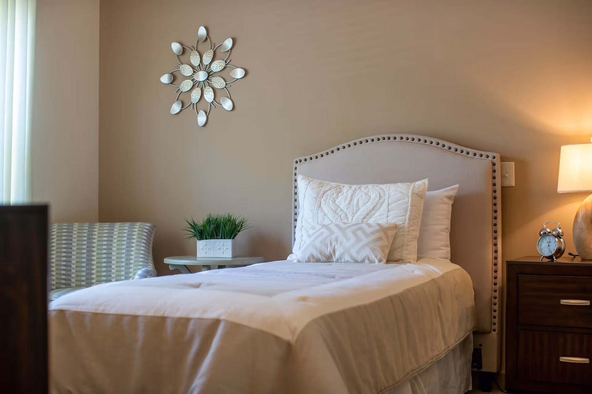 A neatly made single bed with white bedding and decorative pillows in a bedroom. Next to the bed is a wooden nightstand with a lamp and a silver alarm clock. A small round table with a potted plant and a patterned armchair are visible in the background. A decorative wall piece hangs above the bed on a beige wall.