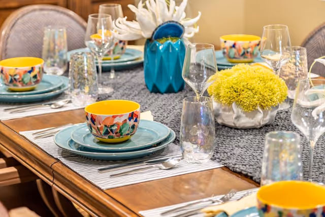 A wooden dining table set with colorful floral bowls, blue plates, clear drinking glasses, wine glasses, and silverware. The table has a gray textured runner with two decorative centerpieces: a blue vase with white coral-like decoration and a white bowl with yellow-green flowers.