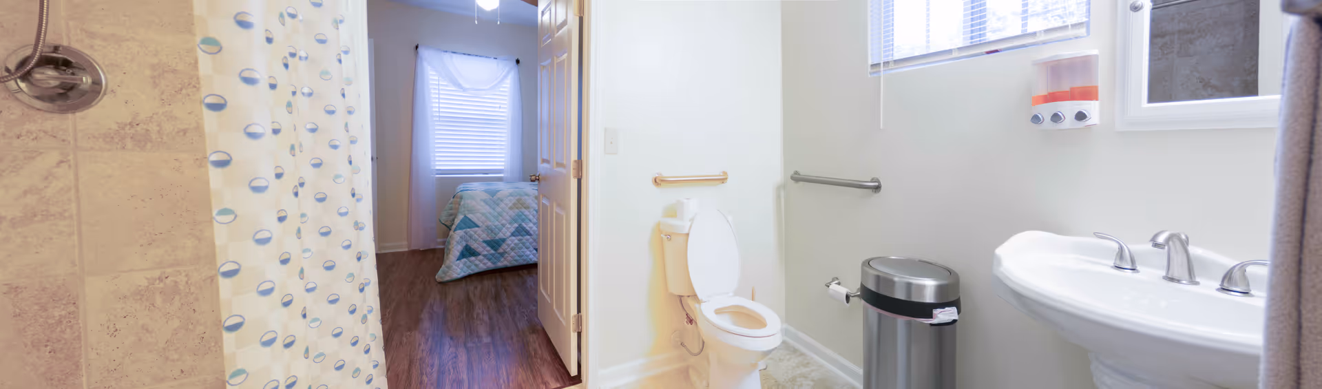 View of a bathroom with a shower curtain on the left, a toilet with grab bars, a trash can, and a sink on the right. Through the open door, a bedroom with a bed covered in a quilt is visible.
