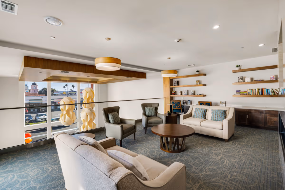 Bright senior living lounge with sofas, armchairs, a round coffee table, built-in shelving and large windows overlooking the street.