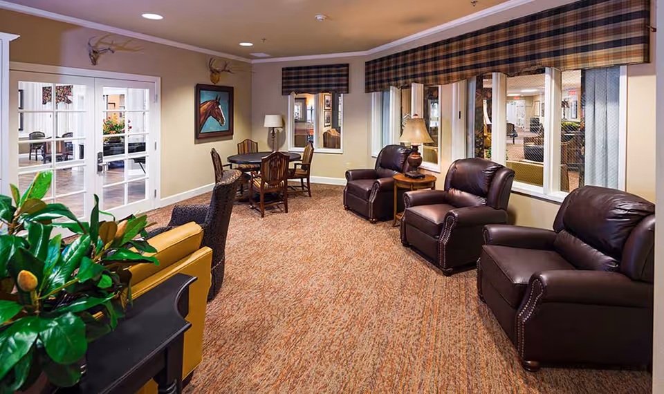 A cozy living room area with three dark brown leather armchairs, a small wooden side table with a lamp, a round table with four chairs, and a yellow armchair partially visible. The room has beige walls, carpeted floor, windows with plaid valances, and decorative elements including a painting of a horse and mounted antlers. There are glass double doors leading to another room with more seating.
