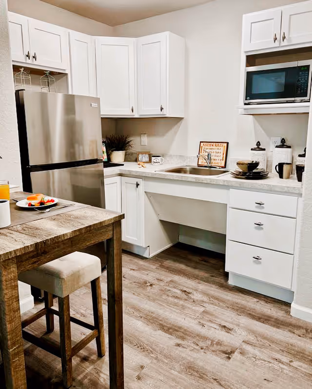 A modern kitchen with white cabinets, a stainless steel refrigerator, a microwave, and a sink. There is a wooden table with a cushioned stool, a plate with food, and a cup on the table. The floor is wood-style laminate, and there are decorative items on the countertop including a small plant and a framed sign.