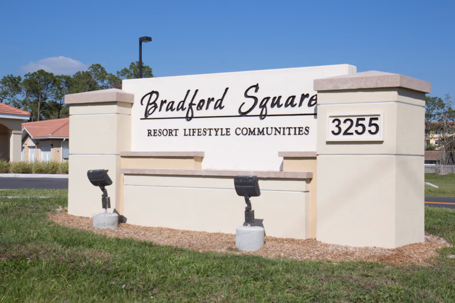 Outdoor entrance sign for Bradford Square Resort Lifestyle Communities with the address number 3255 displayed on a beige stone structure, surrounded by grass and landscaping under a clear blue sky.