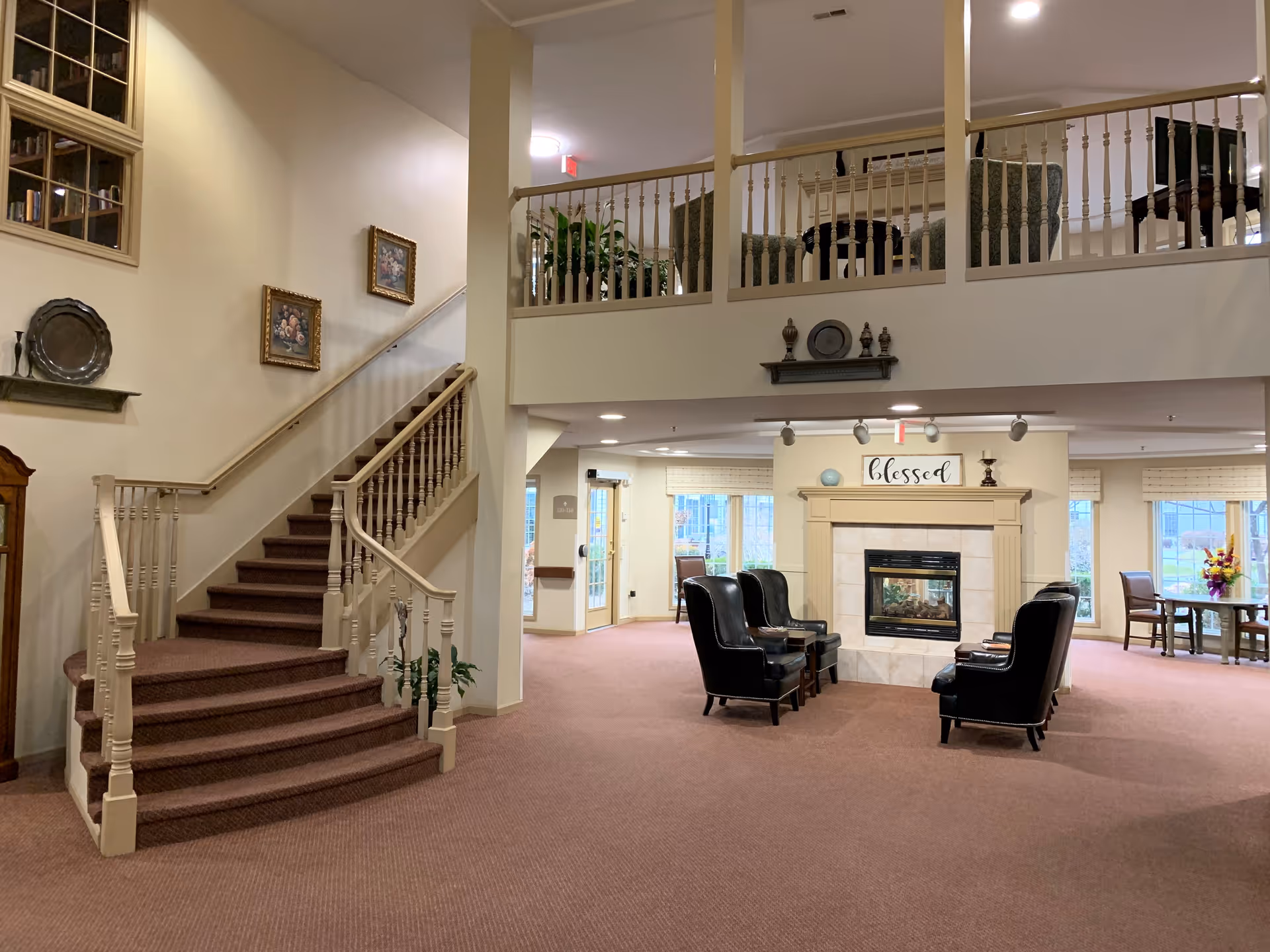 Spacious senior living common area with a carpeted staircase, seating arranged around a central fireplace topped by a 'blessed' sign, and an upper balcony.