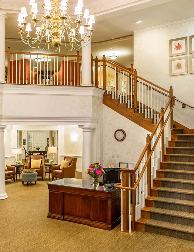 Interior view of a senior living facility lobby with a wooden reception desk adorned with a vase of flowers, a staircase with wooden handrails and carpeted steps leading to an upper floor, a chandelier hanging from the ceiling, and a seating area with chairs and tables in the background.