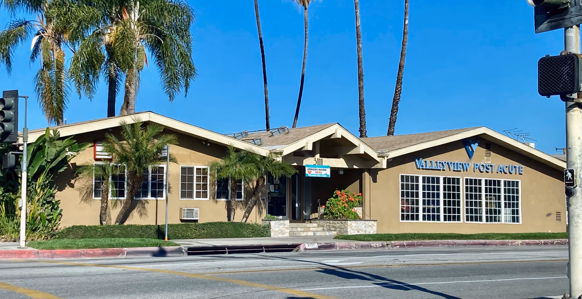 Exterior view of Valley View Post Acute building with beige walls, large windows, and a sign displaying the facility name. Palm trees and other greenery surround the building under a clear blue sky. A pedestrian crossing signal and traffic lights are visible at the street corner in front of the building.