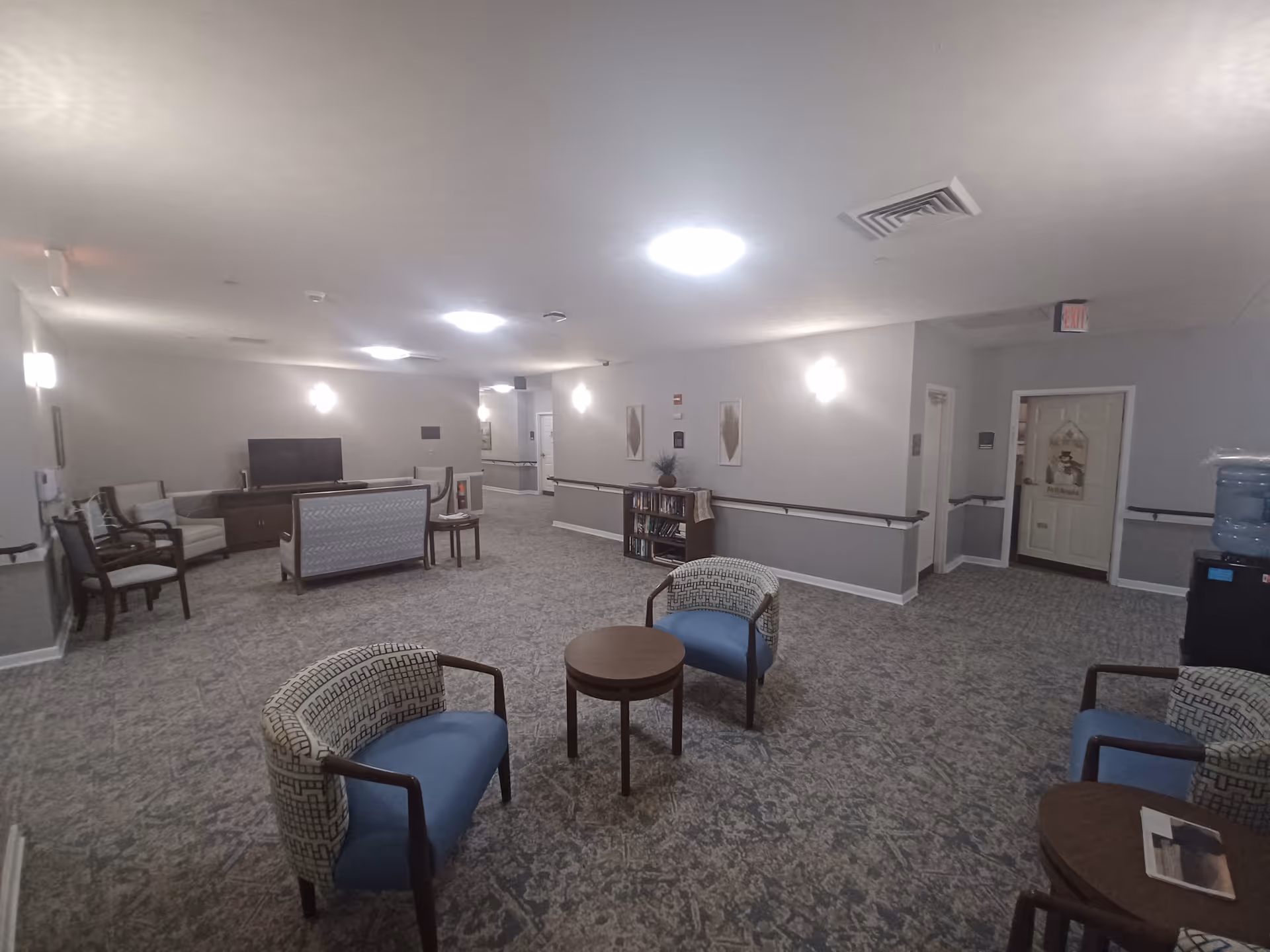 A spacious, well-lit common area in a senior living facility with patterned carpet flooring, several upholstered chairs with blue cushions, small round wooden tables, a television on a wooden stand, and a bookshelf against the wall. The walls are painted light gray with white trim, and there are ceiling lights and wall sconces providing illumination. A water cooler is visible on the right side near a door with a decorative hanging.